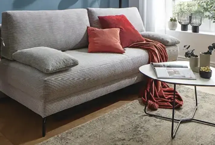 Graues Sofa mit Rillenstoff, roten Kissen und passender Decke, davor runder weisser Couchtisch mit offenem Magazin auf Teppich vor Fensterbank mit Pflanzen.